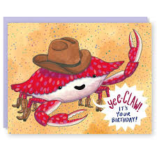 A greeting card atop a periwinkle violet envelope. The front of the card depicts a cute, jolly-looking crab wearing a cowboy hat; on a background of speckled sand. In a large word bubble, the crab exclaims: 