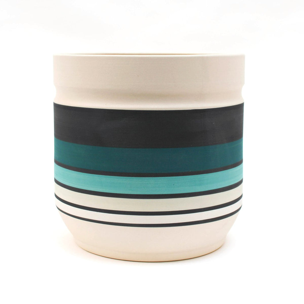 Pacific Planter: a large cylindrical planter painted with horizontal stripes. Each large colored stripe is a different shade of blue, with thinner black stripes in between each colored stripe. From top to bottom, the colors are: white, navy blue, teal, sky blue, light grey, and white.