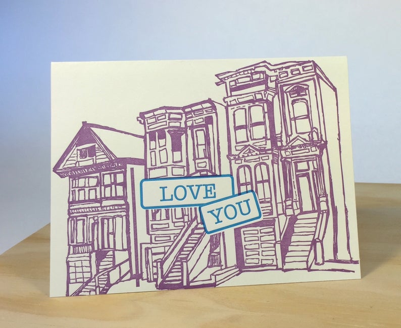 The front of this card depicts a purple line drawing of three Victorian-style houses, which have three stories and have narrow structures. Teal text in two matching teal rectangles in the middle of the card reads: 