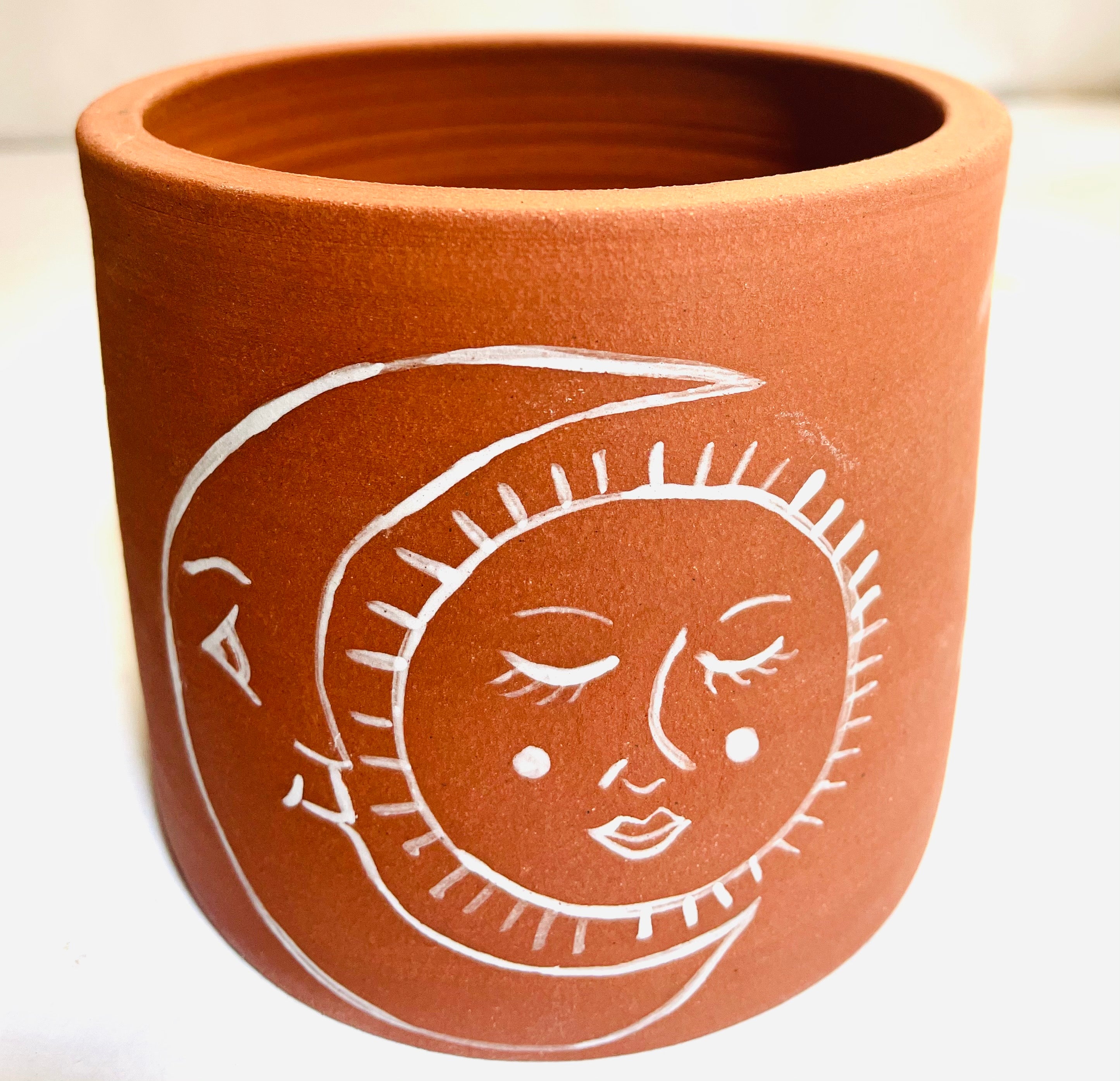 Luna y Sol Planter: an unglazed red clay cylindrical planter with a white handpainted design of a large crescent moon, with a serenely smiling sun in the space between the crescent's points.