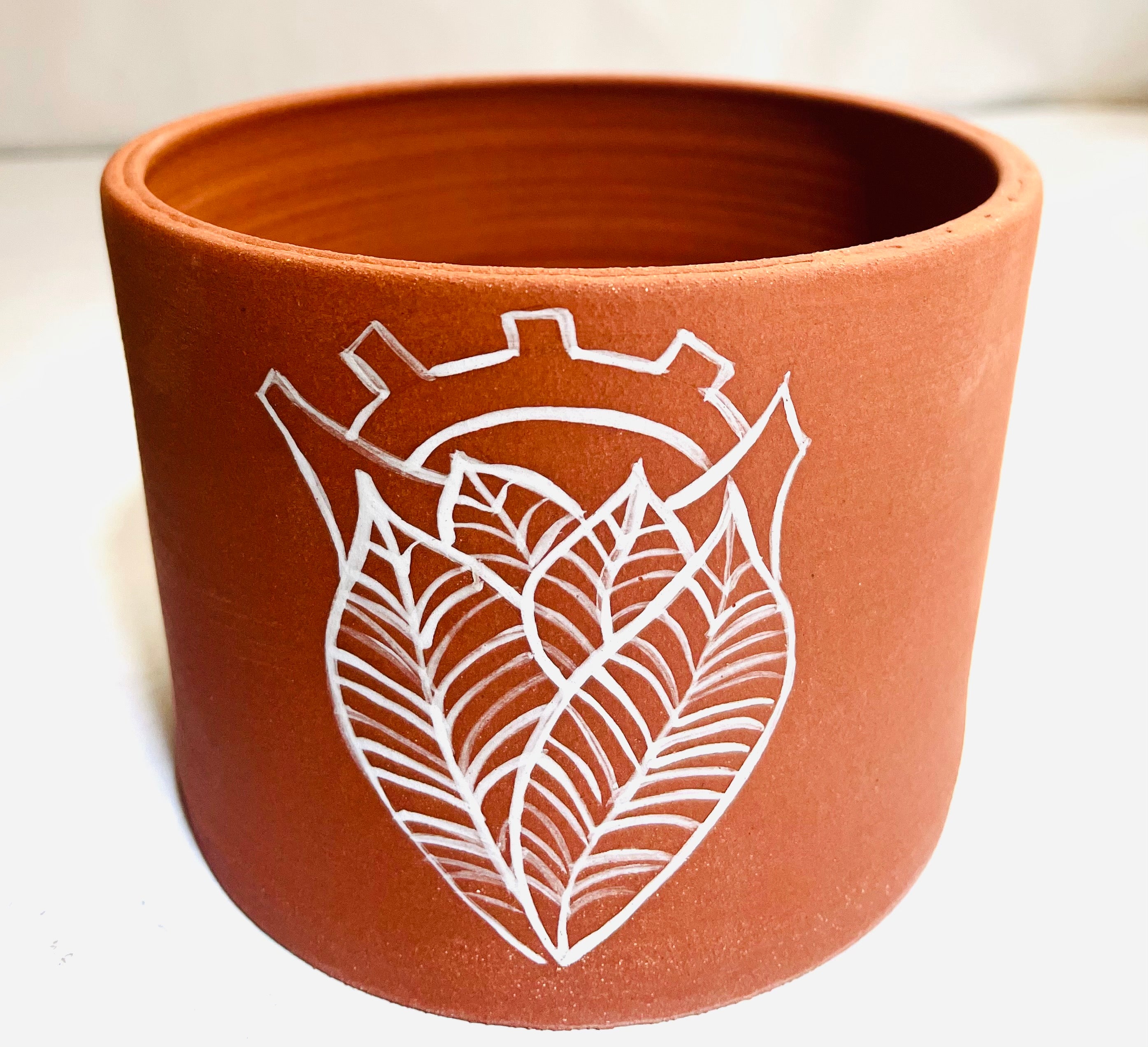 Sacred Heart Red Clay Planter: A red, unglazed cylindrical planter with a white handpainted design of an anatomical sacred heart. The heart is made of veined leaves.