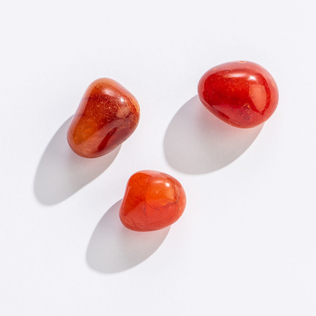 Tumbled Carnelian: three small, smooth, rounded stones on a white background. The stones vary in hue but are generally deep red with orange tones.