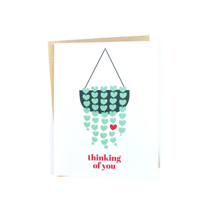 The front of this card depicts a hanging planter, which has a long lush trailing String of Hearts plant growing out of it. All of the plant's leaves are green, with the exception of one which is red. Matching red text underneath the planter reads: 